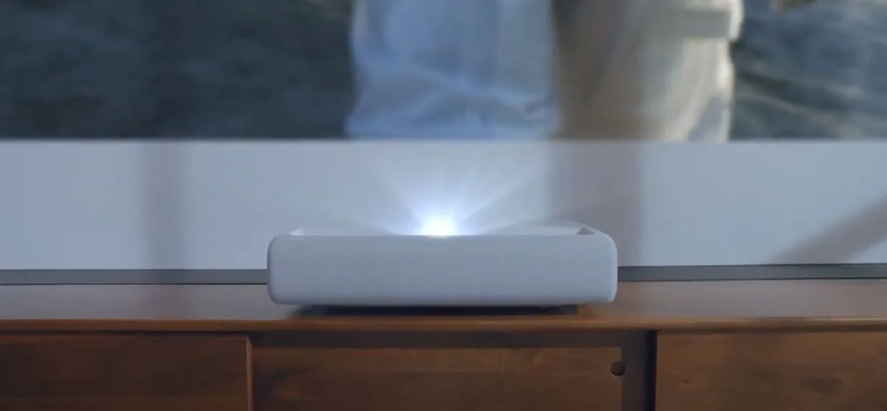 Samsung The Premiere LSP9T ultra-short-throw projector on a wooden sideboard, white light flaring from the lens on its top surface