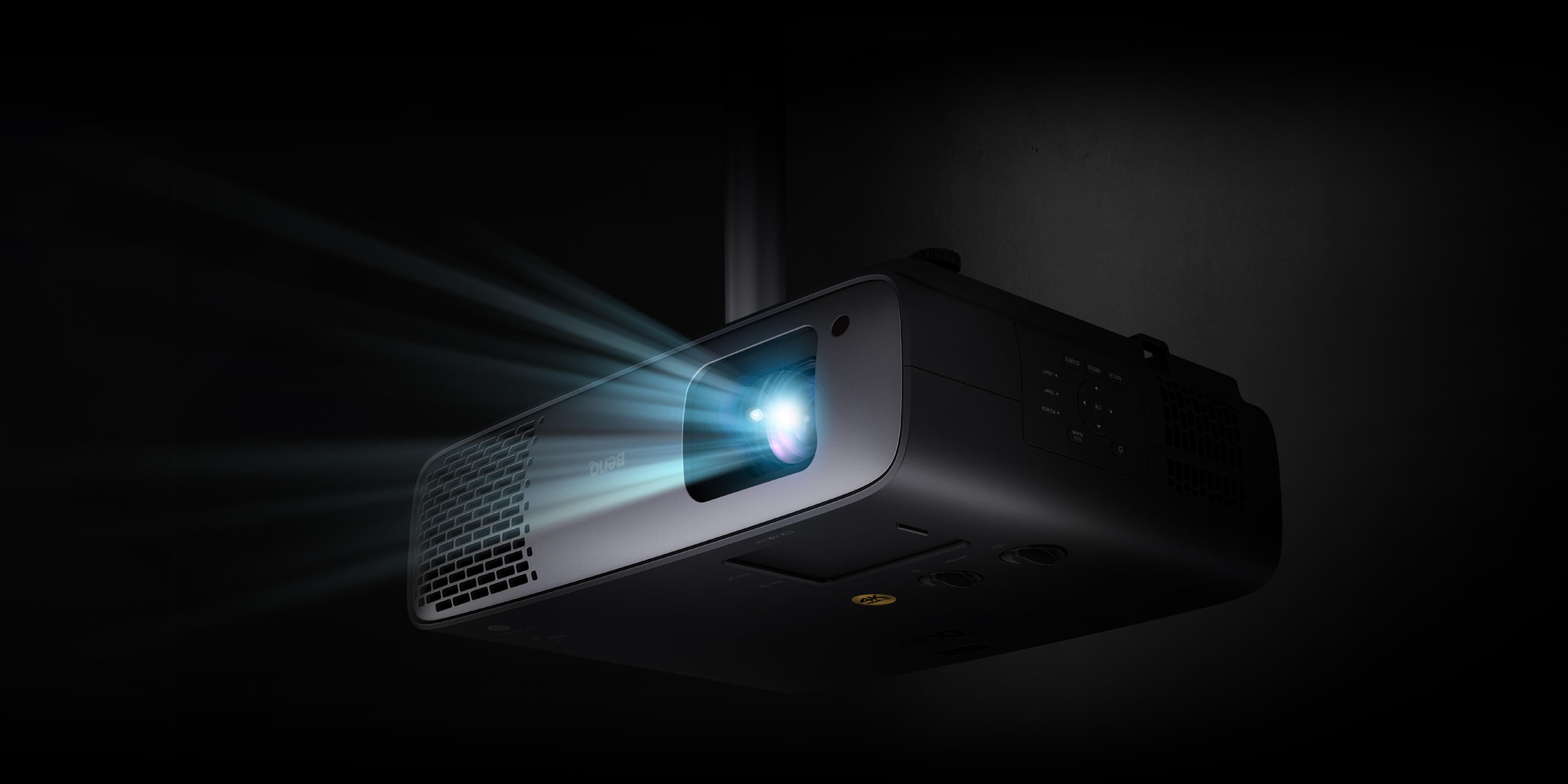 BenQ W4100i seen from below at a three-quarter angle, ceiling mounted, with a beam of light leaving the lens in a dark room