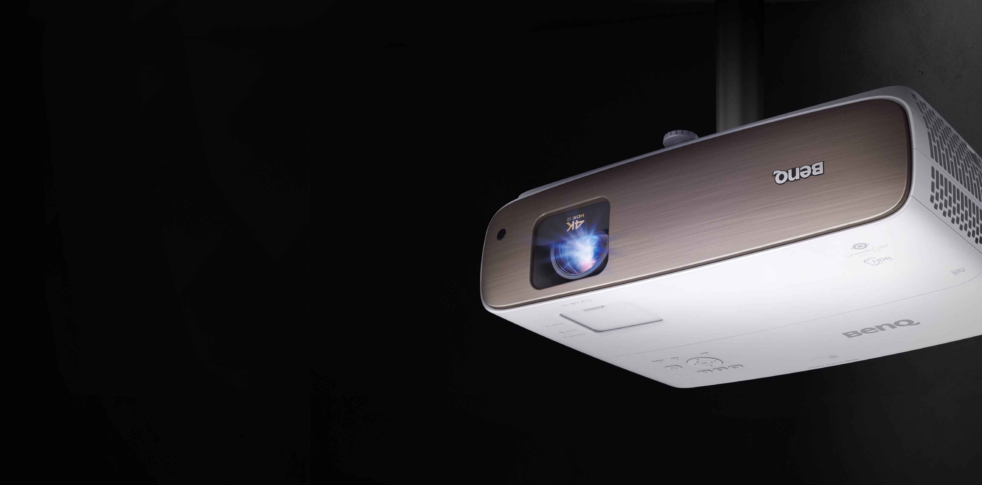 BenQ HT3560 projector ceiling-mounted upside down, front three-quarter view with a blue glow from the lens against a dark background