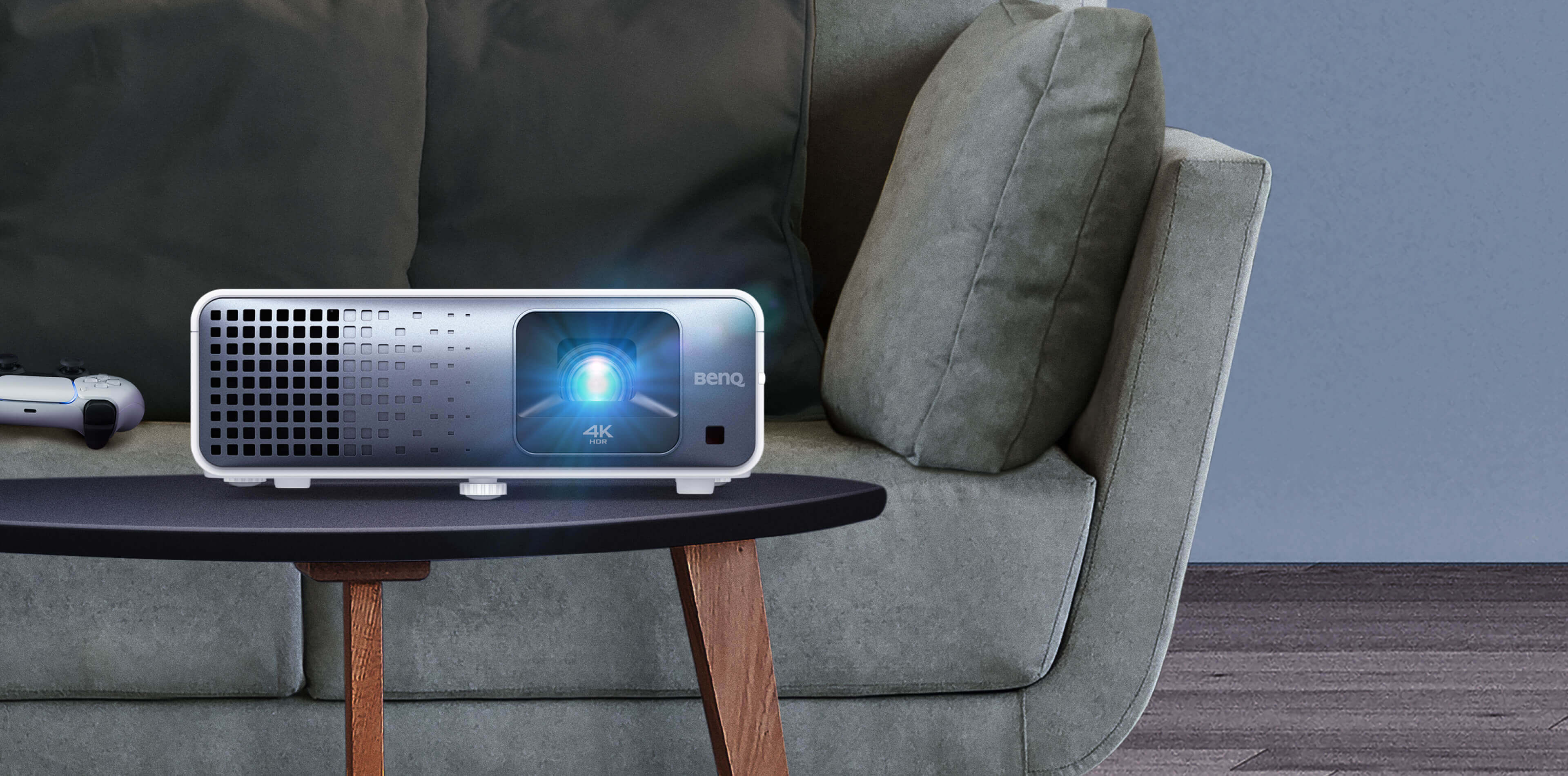 BenQ TK710 projector on a coffee table in front of a sofa, lens glowing, with a game controller beside it