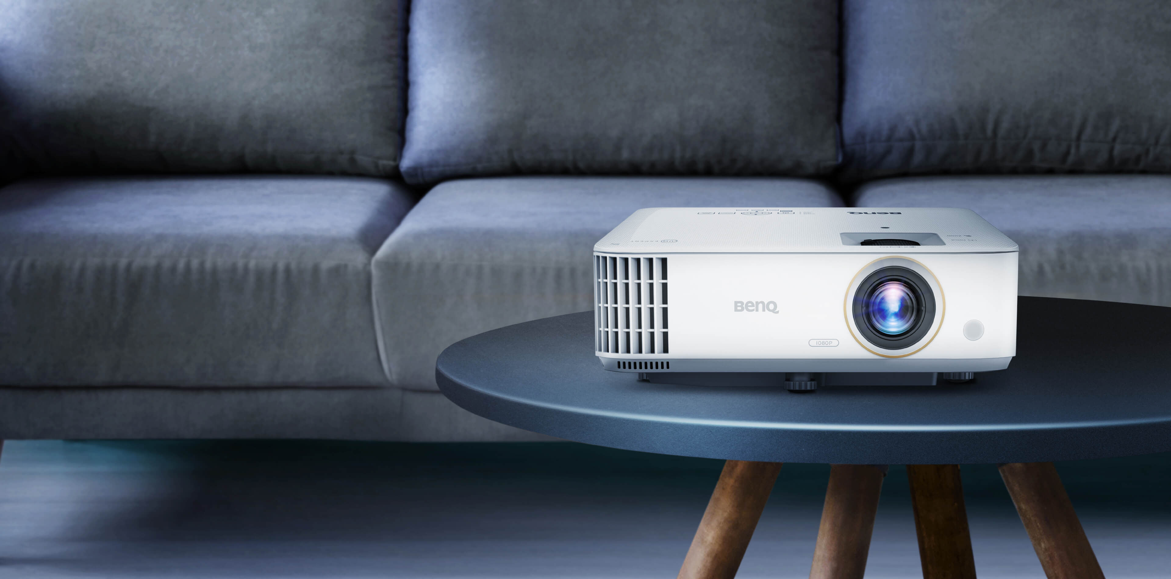 BenQ TH685i white projector on a round table in front of a sofa, seen from the front three-quarter angle