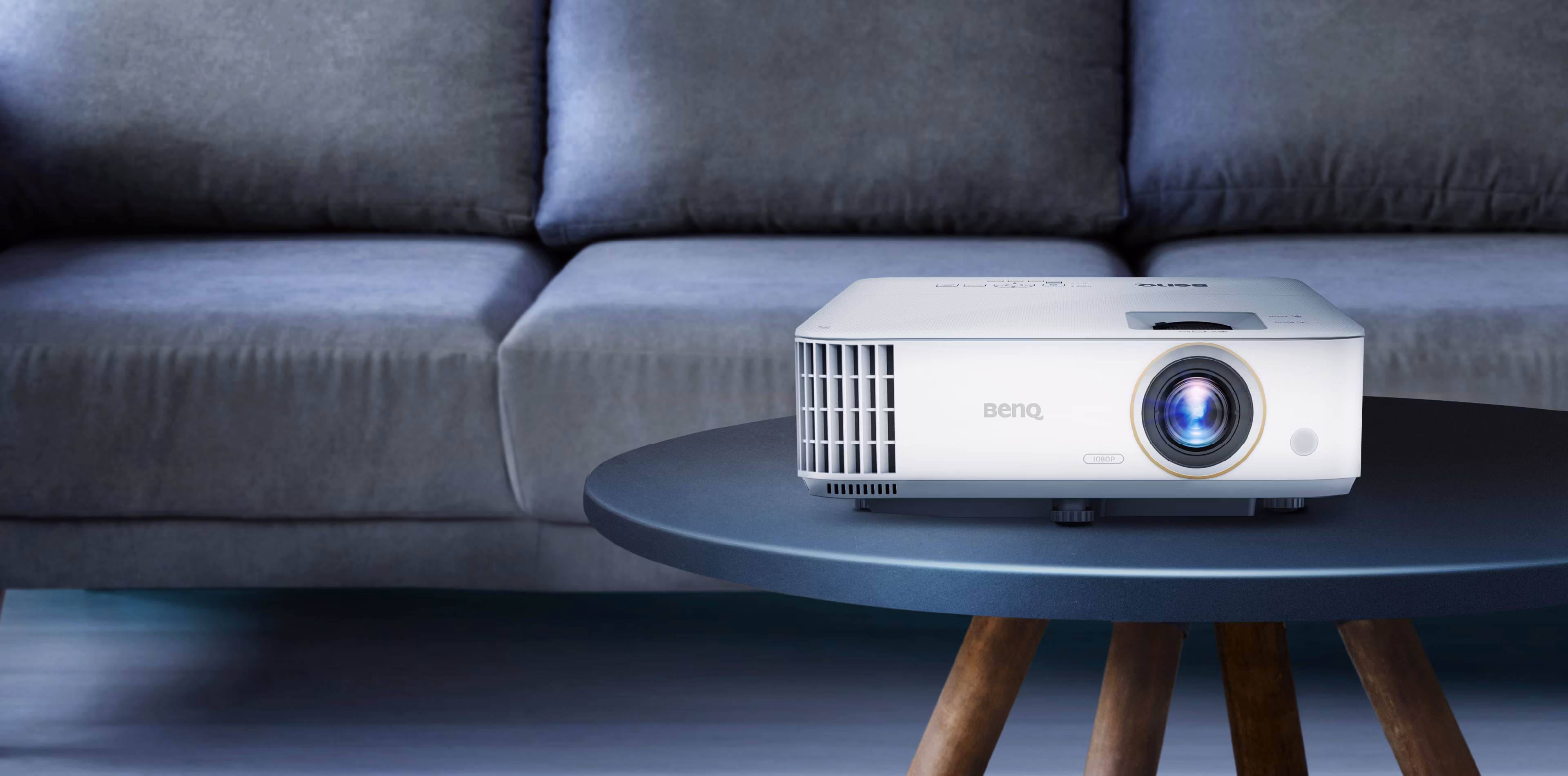 BenQ TH585 white projector with a gold-ringed lens on a round side table in front of a grey sofa